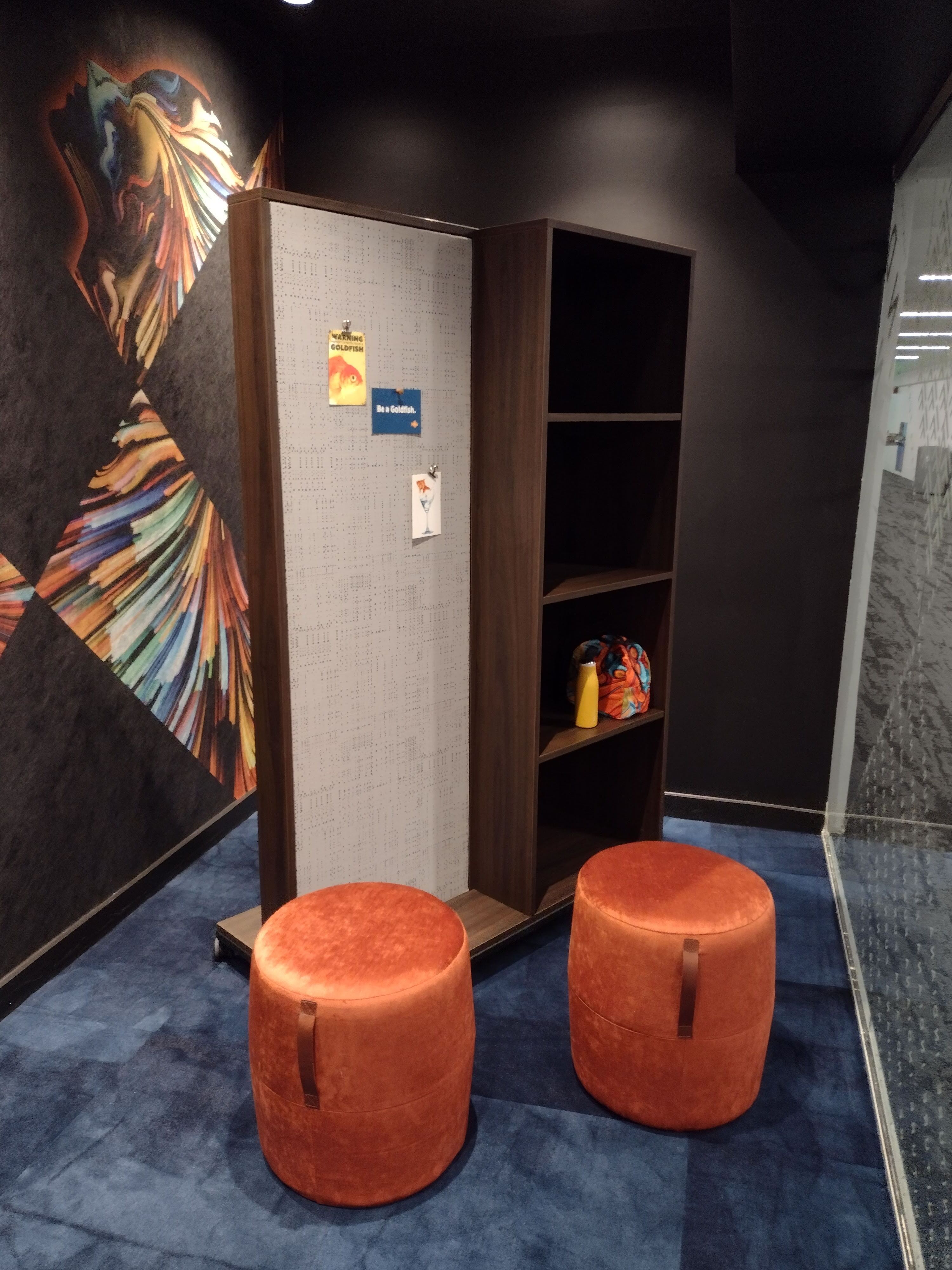 An image of two cylindrical orange stools next to a moveable divider that has storage cubbies and some soundproofing fabric. 